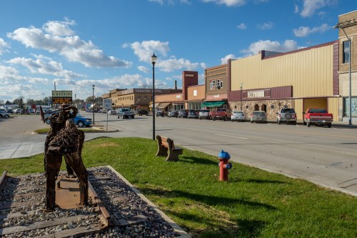 A street view of a small town with shops, cars, and a sculpture near the sidewalk under a blue sky with clouds.