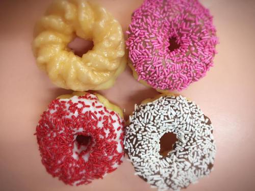 Four colorful donuts: one yellow, one pink with sprinkles, one white with sprinkles, and one red with sprinkles.