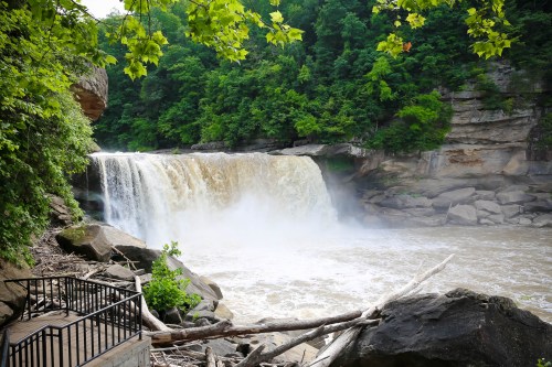 A waterfall cascades over rocks, surrounded by lush green trees and a wooden railing in the foreground.