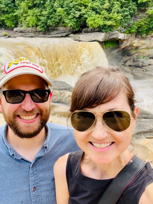 A couple smiles for a selfie in front of a waterfall, surrounded by lush greenery.