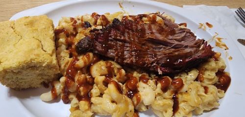 A plate of macaroni and cheese topped with barbecue beef, alongside a piece of cornbread.
