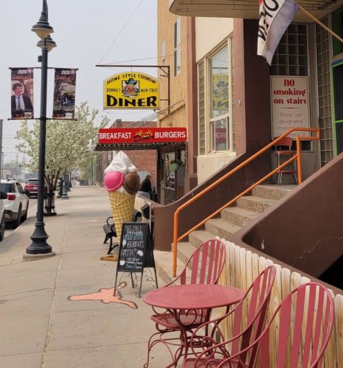 A diner with a large ice cream cone sign outside, featuring pink and brown scoops, and red chairs on the sidewalk.
