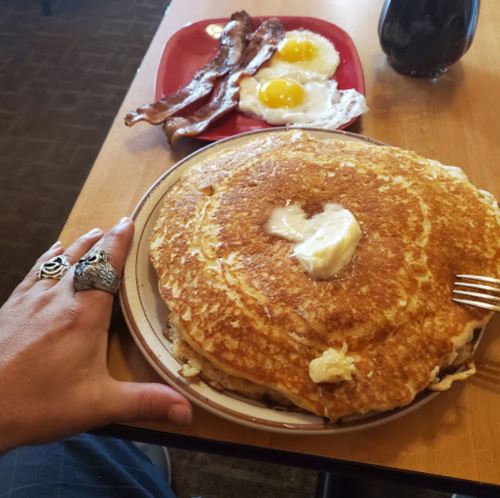A hand with rings reaches for a large stack of pancakes topped with butter, alongside a plate of bacon and eggs.