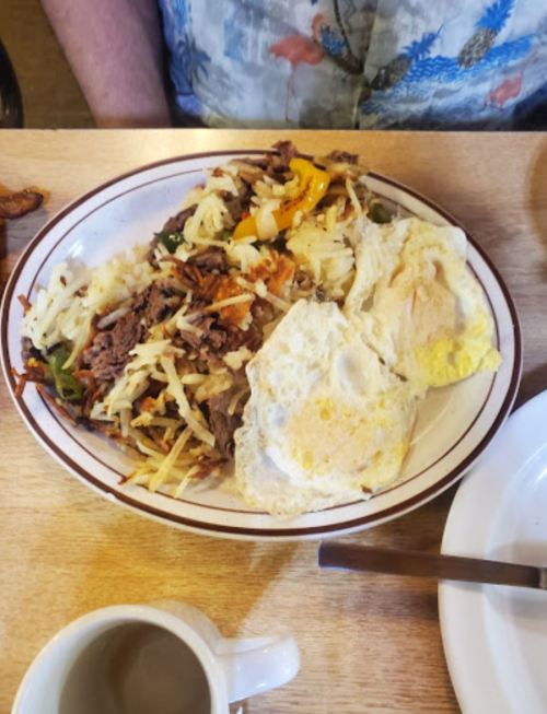 A plate of hash browns topped with beef, peppers, and two fried eggs, served with a cup of coffee on the side.