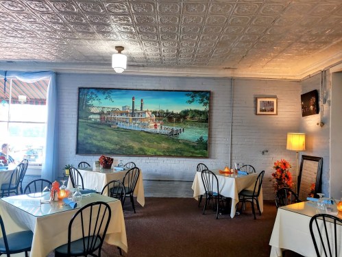Cozy restaurant interior with tables set for dining, featuring a large painting of a steamboat on the wall.
