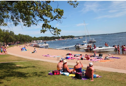 A sunny beach scene with people relaxing on the sand, boats in the water, and trees lining the shore.