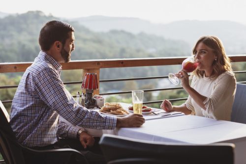 A couple enjoys a meal on a balcony with scenic views, sipping drinks and sharing a moment together.
