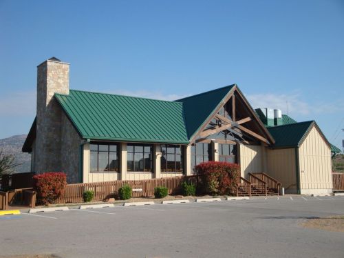 A modern building with a green metal roof, large windows, and a stone chimney, surrounded by landscaping.