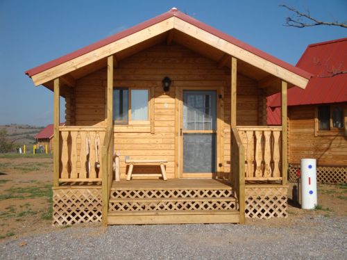 A small wooden cabin with a red roof, featuring a porch and a gravel path in a rural setting.