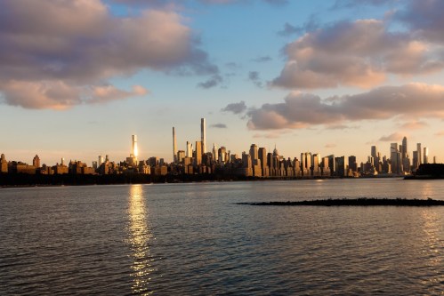 City skyline at sunset, reflecting on water with clouds in the sky and tall buildings silhouetted against the horizon.