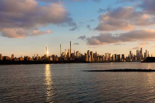 City skyline at sunset, reflecting on water with clouds in the sky and tall buildings silhouetted against the horizon.