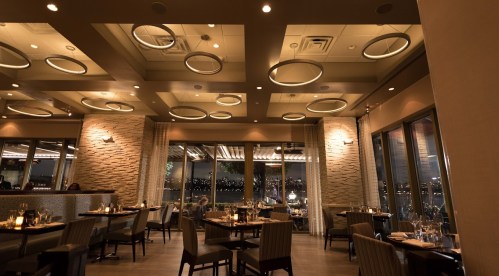 Elegant restaurant interior with modern lighting, tables set for dining, and large windows showcasing a city view.