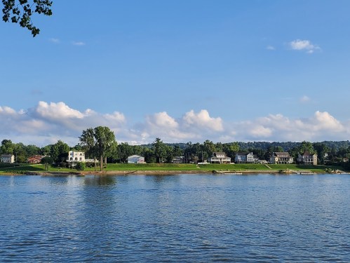 A serene river view with houses lining the shore under a blue sky with fluffy clouds.