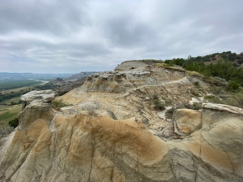A rocky landscape with layered formations under a cloudy sky, overlooking a valley and distant hills.