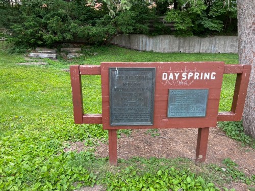 Sign for "Day Spring" with historical plaques, surrounded by greenery and a stone path in the background.