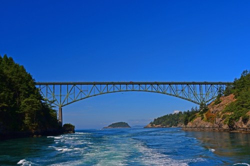 A green bridge spans a blue waterway, surrounded by lush trees and a clear blue sky.