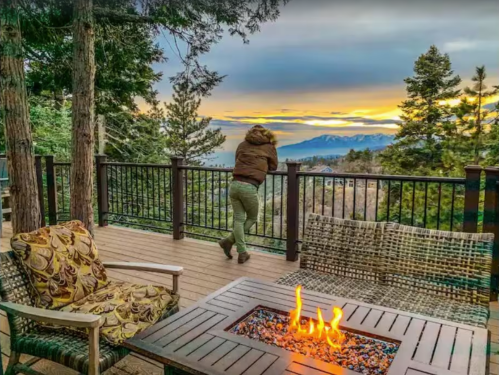 A person leans on a railing, gazing at a sunset over mountains, with a cozy outdoor seating area and fire pit nearby.