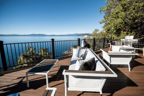 A scenic outdoor patio with modern furniture overlooking a tranquil lake and mountains under a clear blue sky.