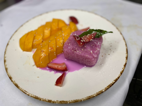 A plate featuring purple sticky rice, mango slices, and strawberries, garnished with a mint leaf and drizzled with sauce.