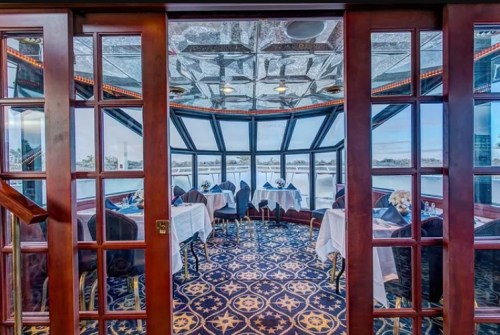 Elegant dining room with large windows overlooking water, featuring white tablecloths and a decorative ceiling.