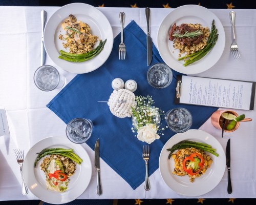 A table set with four plates of gourmet meals, asparagus, water glasses, and a floral centerpiece on a blue napkin.