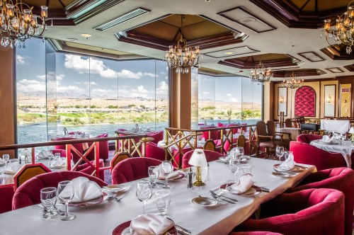Elegant restaurant interior with red seating, chandeliers, and large windows overlooking a scenic view.