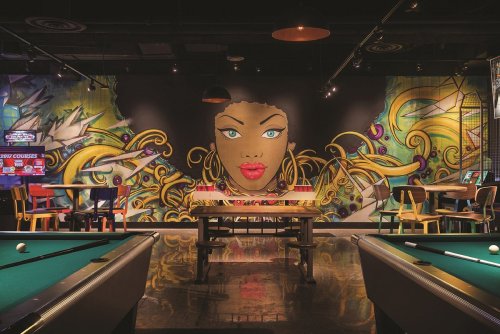 A vibrant mural of a woman's face adorns a game room with pool tables and colorful decor.