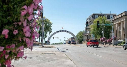 A street view of a town archway with traffic lights, surrounded by greenery and flowers on a sunny day.