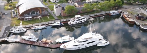 Aerial view of a marina with various yachts and boats docked along a wooden pier, surrounded by greenery and buildings.