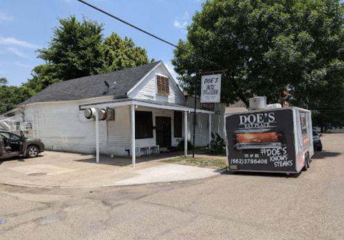 A small white building with a sign for Doe's and a food truck parked outside on a sunny day.