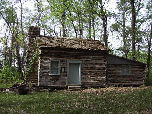 A rustic log cabin surrounded by trees, featuring a stone chimney and a simple front door.