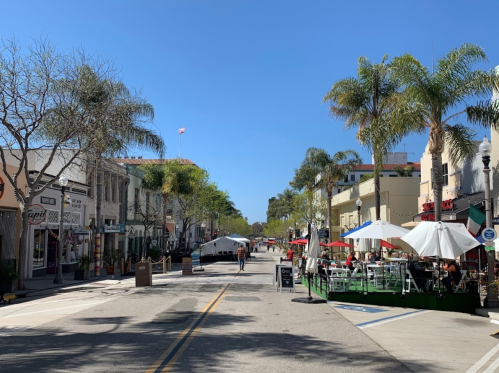 A sunny street lined with shops and outdoor dining, featuring palm trees and a clear blue sky.