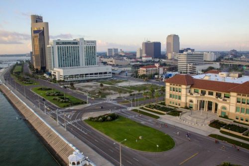 Aerial view of a coastal city with modern buildings, roads, and a waterfront promenade under a clear sky.