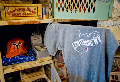 A gray t-shirt with "Lewisburg, WV" printed on it hangs next to a brown hat in a rustic shop display.