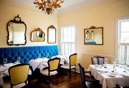 Cozy restaurant interior with blue banquette, elegant mirrors, and white tablecloths set for dining.