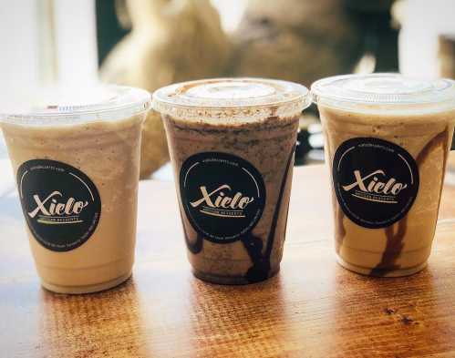 Three cups of blended desserts with lids, featuring different flavors and topped with chocolate and caramel drizzles.