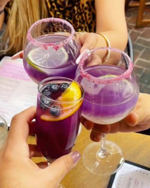 Three colorful cocktails are clinked together, featuring purple drinks with fruit garnishes and decorative rims.
