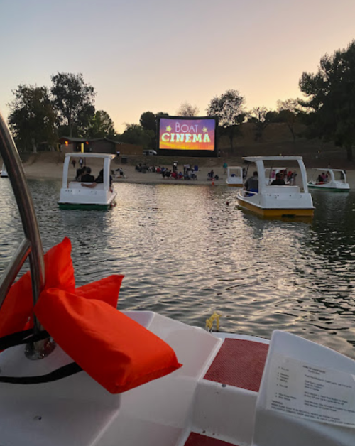 A view from a boat showing a large screen labeled "Boat Cinema" with people gathered on the shore at sunset.