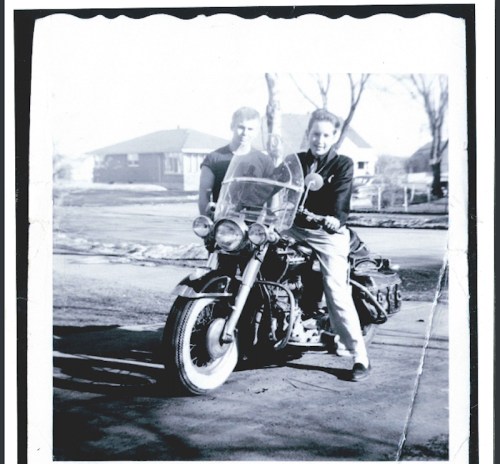 Two young men pose with a vintage motorcycle in a black-and-white photo, with houses visible in the background.