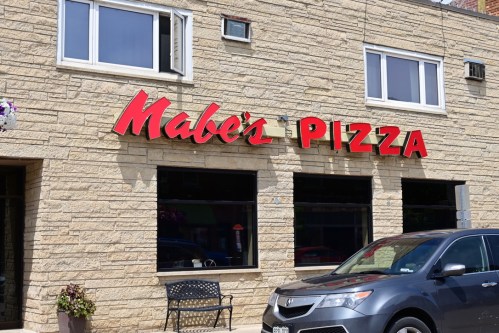 Sign for Mabe's Pizza on a stone building, with a parked car and a bench in front.