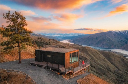 Modern cabin on a hillside with a stunning sunset view over mountains and a river.