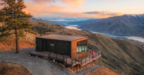 A modern cabin on a hillside with a scenic view of mountains and a river at sunset. A person stands on the deck.