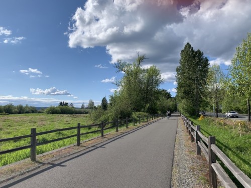 A scenic pathway lined with trees and a wooden fence, under a blue sky with clouds, ideal for walking or biking.