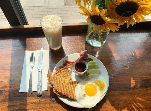 A plate of breakfast with eggs, bacon, toast, and fruit, alongside a glass of iced coffee and a vase of sunflowers.