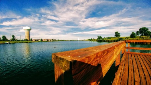 A serene lake view with a wooden dock, lush greenery, and a water tower in the background under a blue sky.