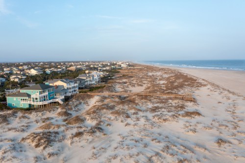 Aerial view of a sandy beach with houses along the shore and a wooden walkway leading to the water.