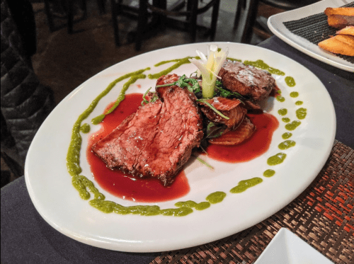 A plate of sliced beef with a red sauce, garnished with greens and drizzled with green sauce, served on a white dish.