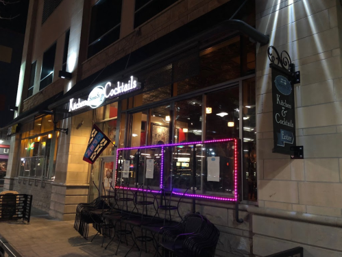 Exterior of a restaurant named "Kitchen & Cocktails," featuring neon lights and outdoor seating at night.