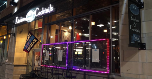 Exterior of a restaurant with neon lights, featuring large windows and a sign that reads "Kitchen & Cocktails."