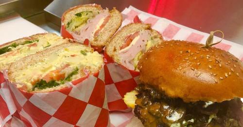 A sesame seed bun burger and a sliced sandwich with layers of meat, lettuce, and tomato, served on checkered paper.